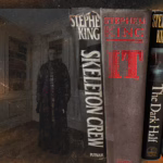 A surreal and eerie digital collage featuring five Stephen King novels prominently aligned in a vertical row: "Skeleton Crew," "IT," "The Dark Half," "Four Past Midnight," and part of a sixth partially obscured book. All book spines are shown in vivid detail, with aged textures and wear visible on the covers. Behind the books, semi-transparent and ghostlike, stands a sinister figure in a dimly lit, ornate library room with paneled walls and a bookshelf. The figure is male, pale-faced, wearing a long dark coat and staring blankly forward, adding a haunting presence. The background is overlaid with smoky, gritty textures and orange fiery light leaks, giving the image an ominous, horror-themed atmosphere.